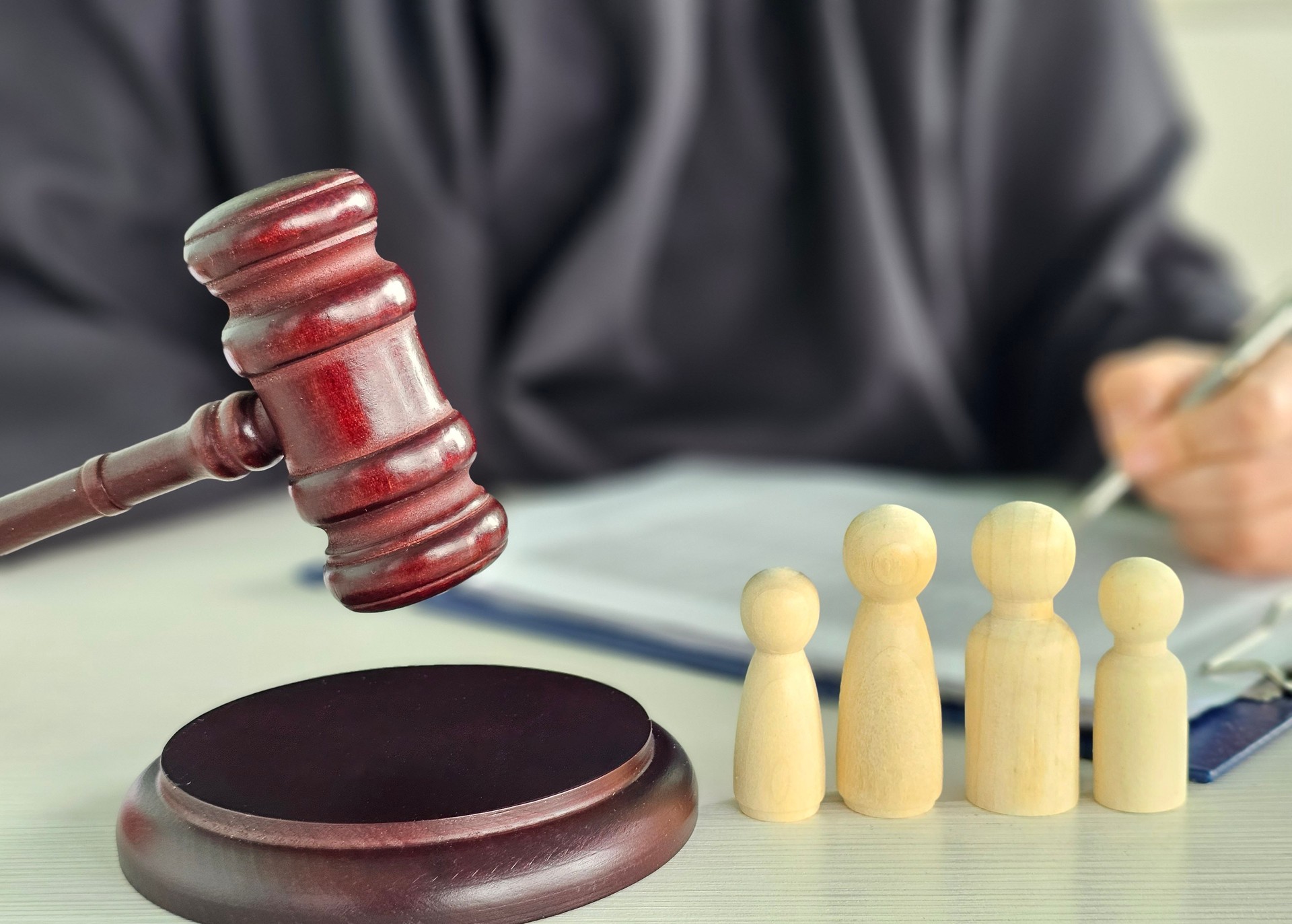 Gavel, sound block and little wooden figures of parents and children placed on desk in courthouse up close, judge and scales of justice in background. Family law, court, divorce, child custody